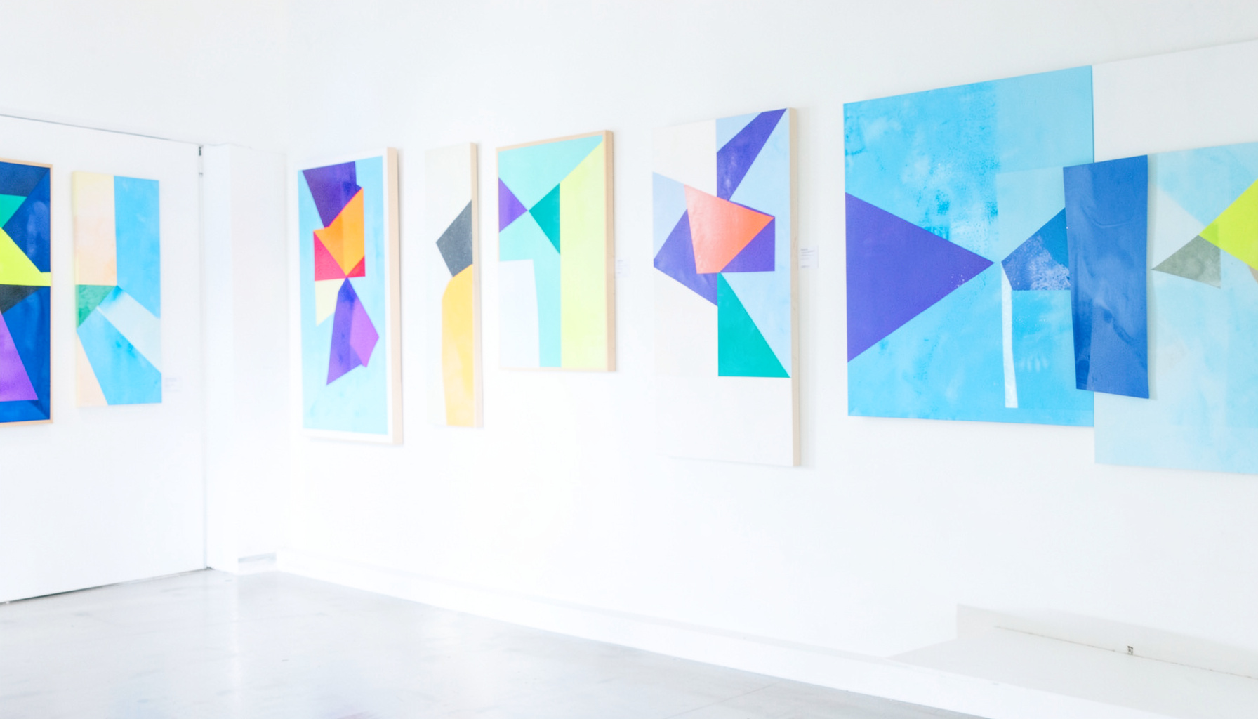 A bright gallery wall with abstract geometric paintings in bold primary colors, seen from a slight angle in soft daylight